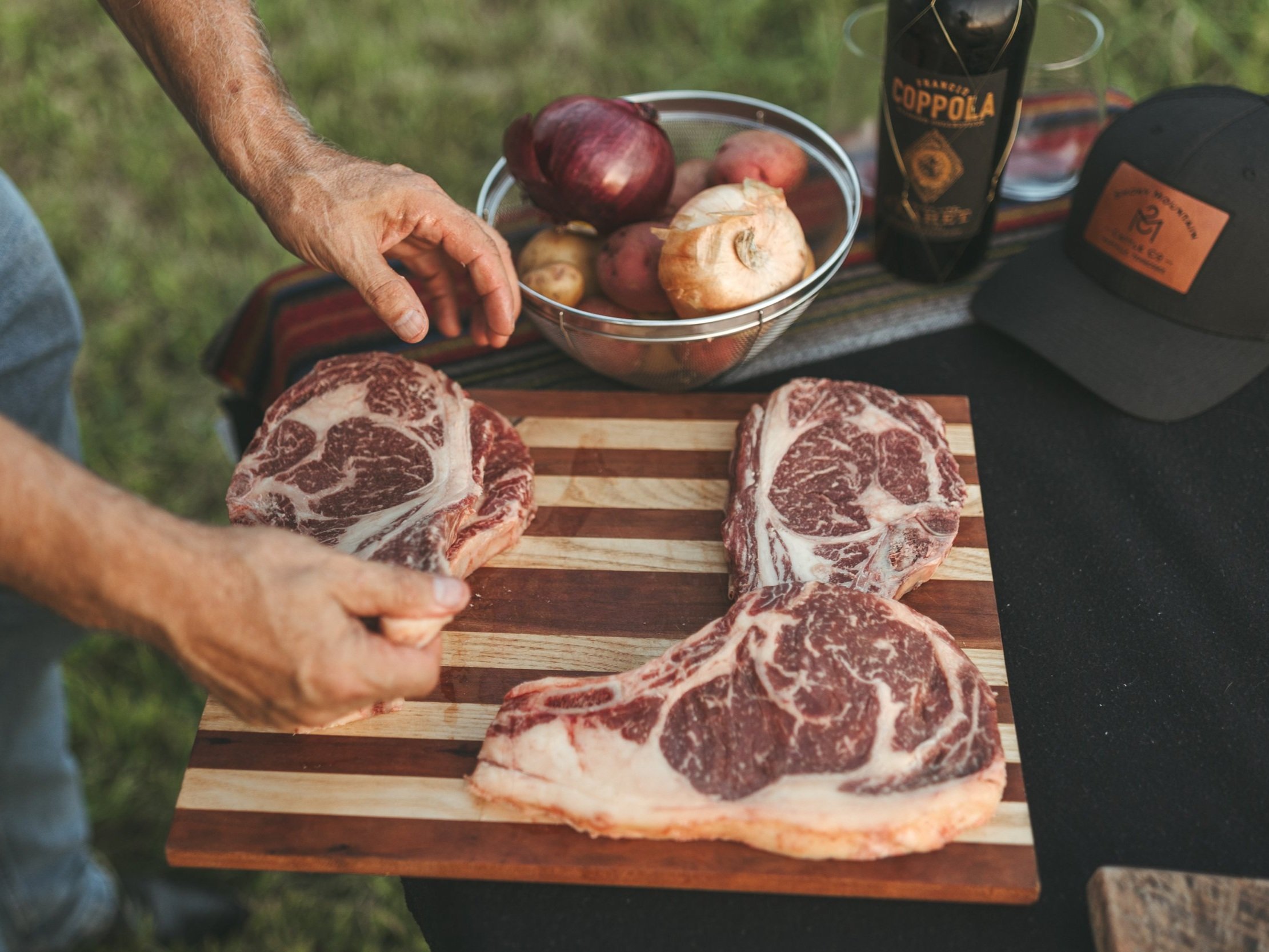 Close-up of a person preparing marbled ribeye steaks on a wooden cutting board, surrounded by fresh vegetables, including red onions and potatoes, and a bottle of wine, set against a grassy outdoor backdrop.