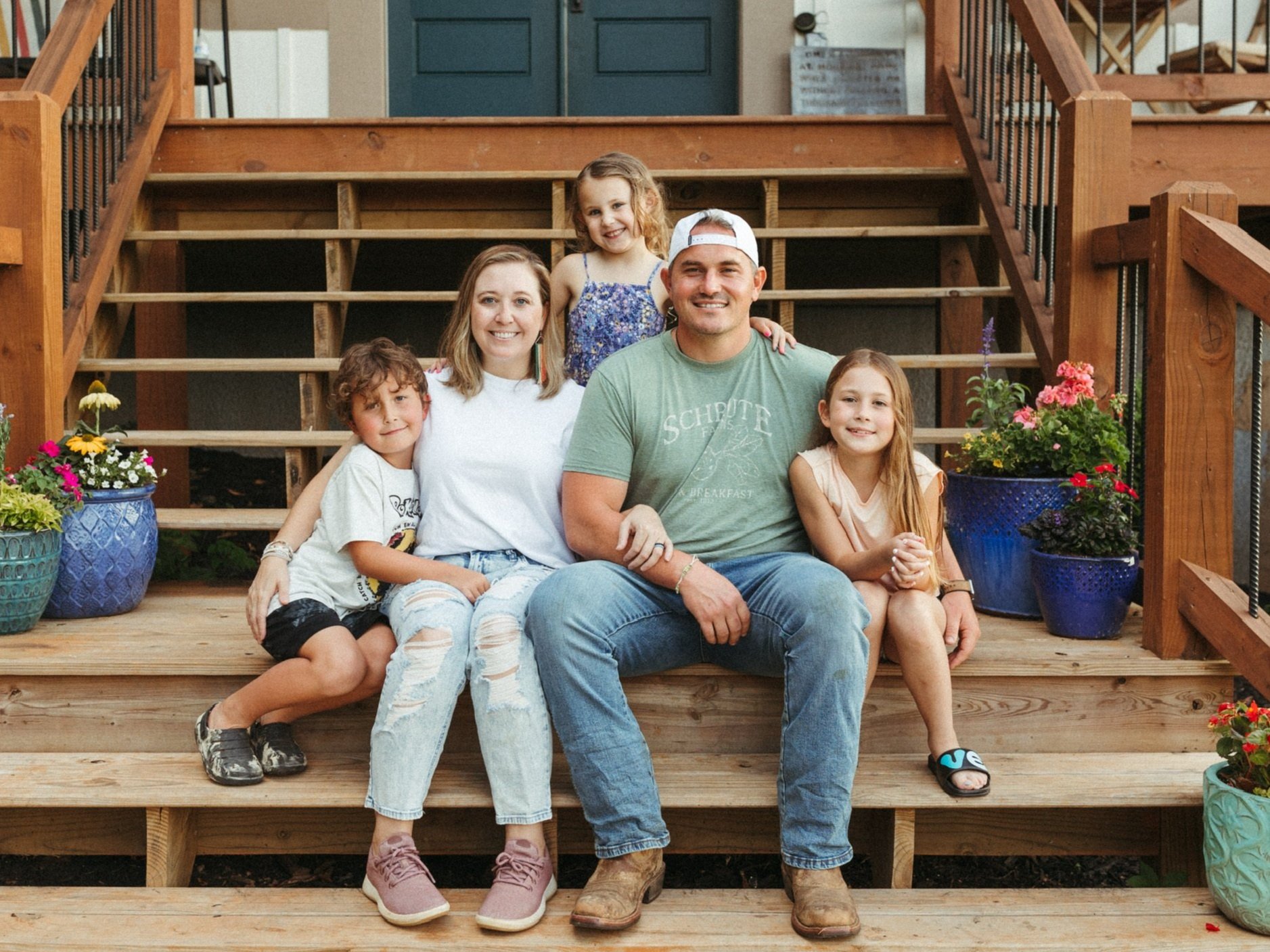 Family portrait on wooden steps, featuring a mother and father with three children. The scene includes vibrant flower pots and a welcoming home entrance in the background. The family is smiling and dressed casually, creating a warm, inviting atmosphere.